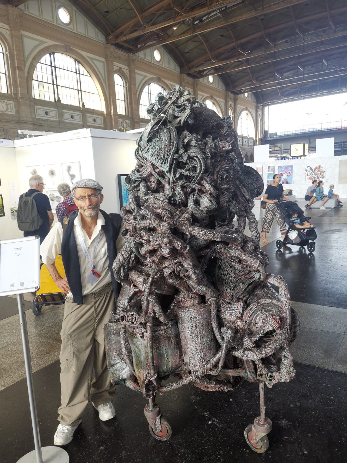 Hohe braun-graue Metallskulptur auf Rollen in einer grossen Ausstellungshalle mit Bogenfenstern und Holzdach; daneben steht ein Mann mit Schiebermütze, im Hintergrund Besucher