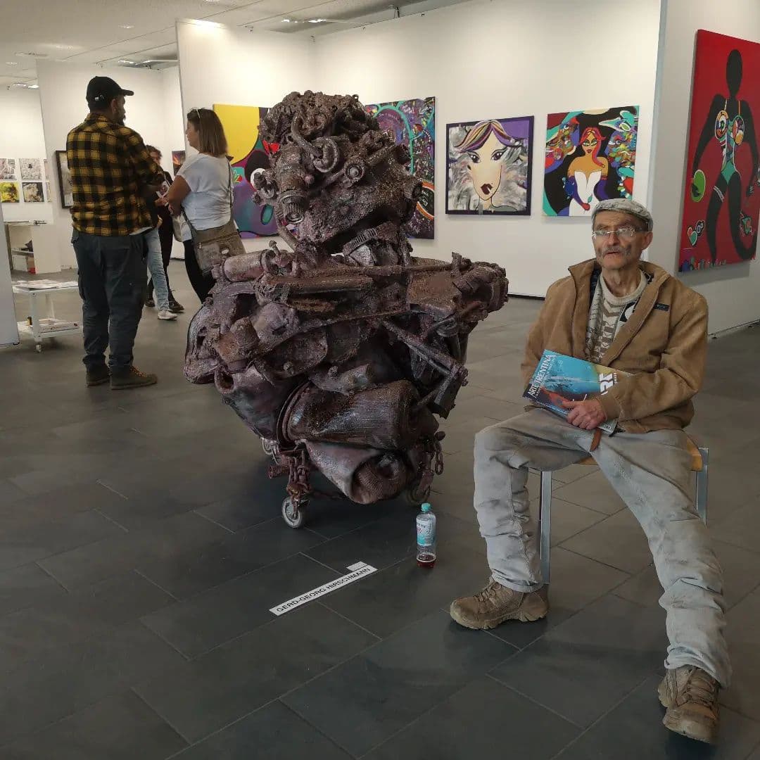 Braune Metallskulptur auf Rollen in einer weissen Messehalle, daneben sitzt ein Mann mit Schiebermütze und einem Heft; an den Wänden Bilder anderer Künstler