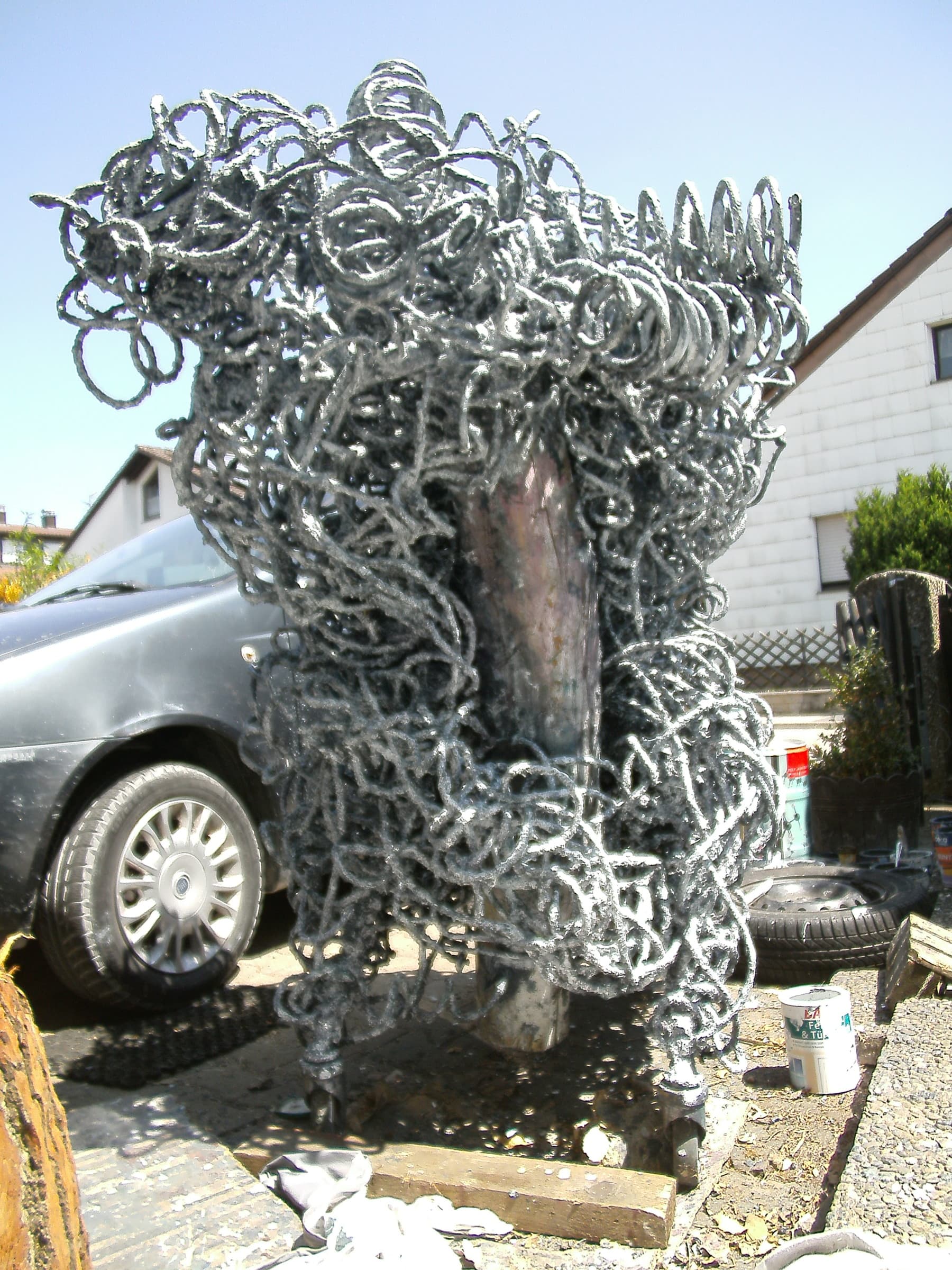 Hohe silbergraue Schlingen-Skulptur mit kupfernem Oval in der Mitte, in einem Hof vor Haus und Auto