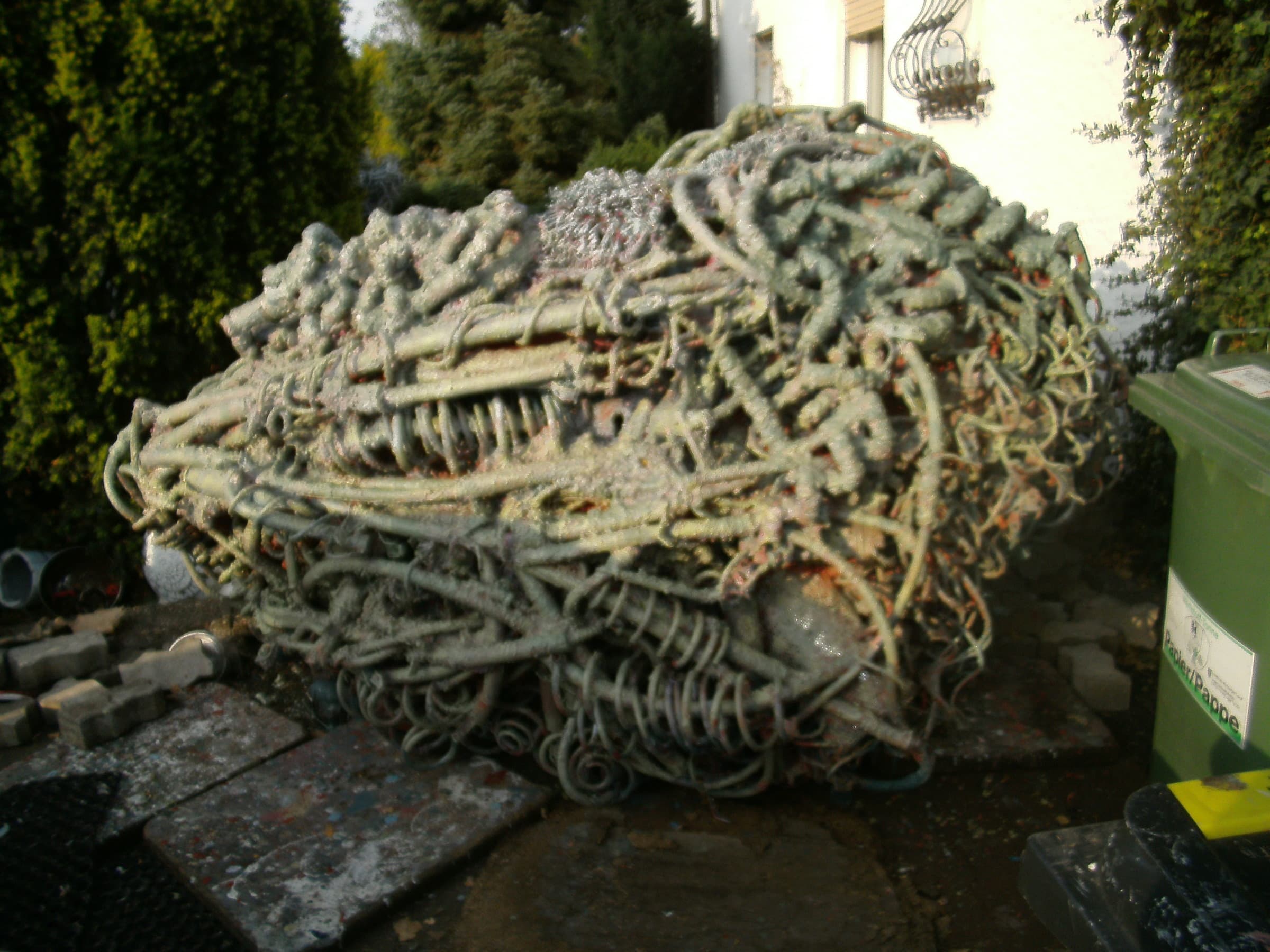 Liegende grün-graue Schlingen-Skulptur in einem Hof neben Hecke und Mülltonne