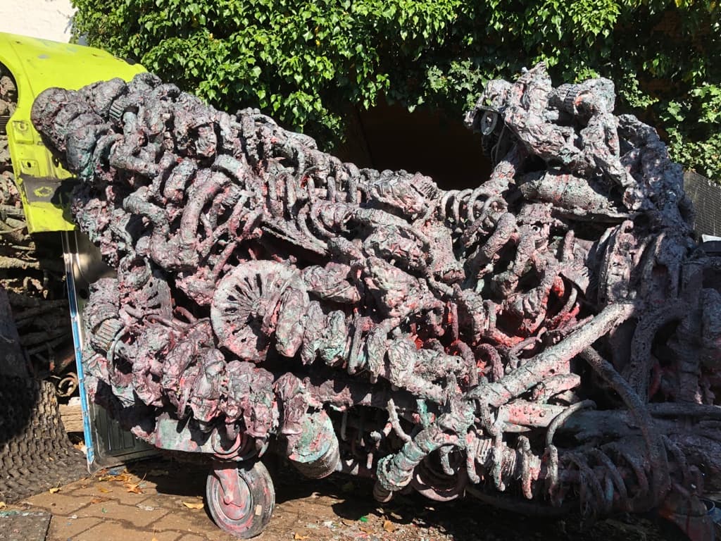 Lange, wagenartige Metallskulptur auf Rollen, violett-rot-grün patiniert, voller Zahnräder und Metallteile, vor einer Hecke