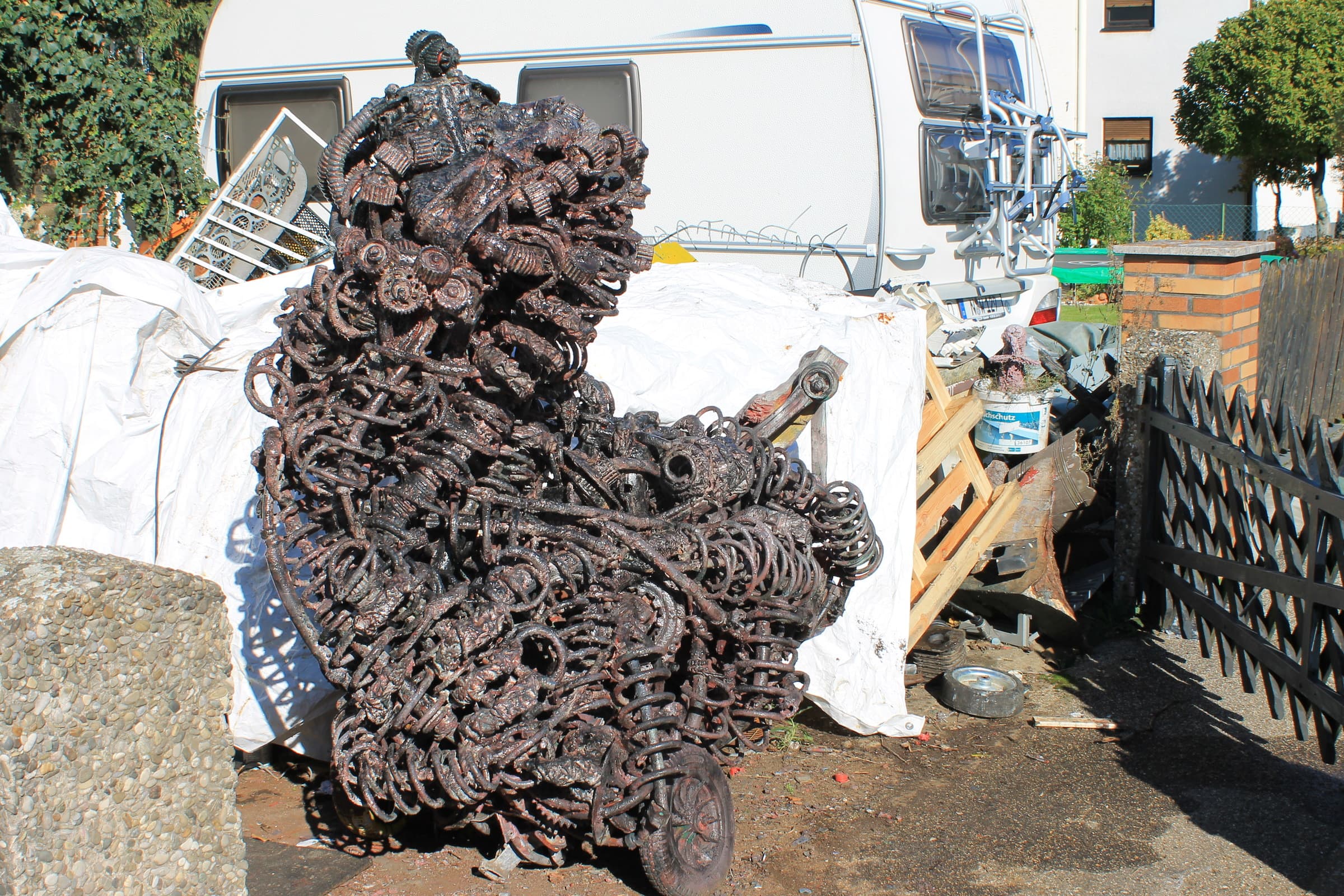 Kupferrot-rostige Metallskulptur aus Federn, Zahnrädern und Metallteilen vor weissen Planen und einem Wohnwagen