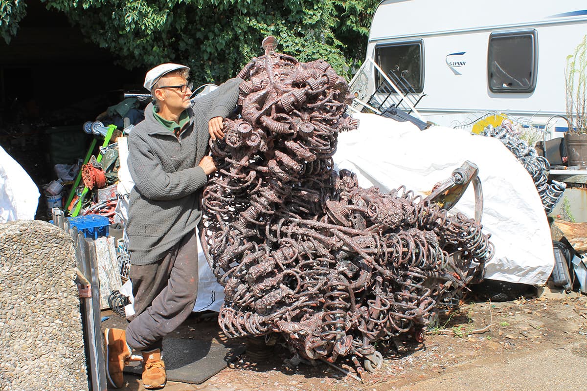 Gerd Georg Hirschmann lehnt an einer kupferroten Metallskulptur aus Federn und Zahnrädern, dahinter ein Wohnwagen und weisse Planen