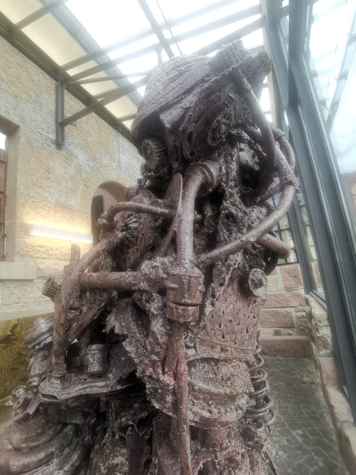 Dunkle Metallskulptur aus Rohren, Gittern und Zahnrädern in einer Glashalle vor einer Sandsteinwand, von unten gesehen