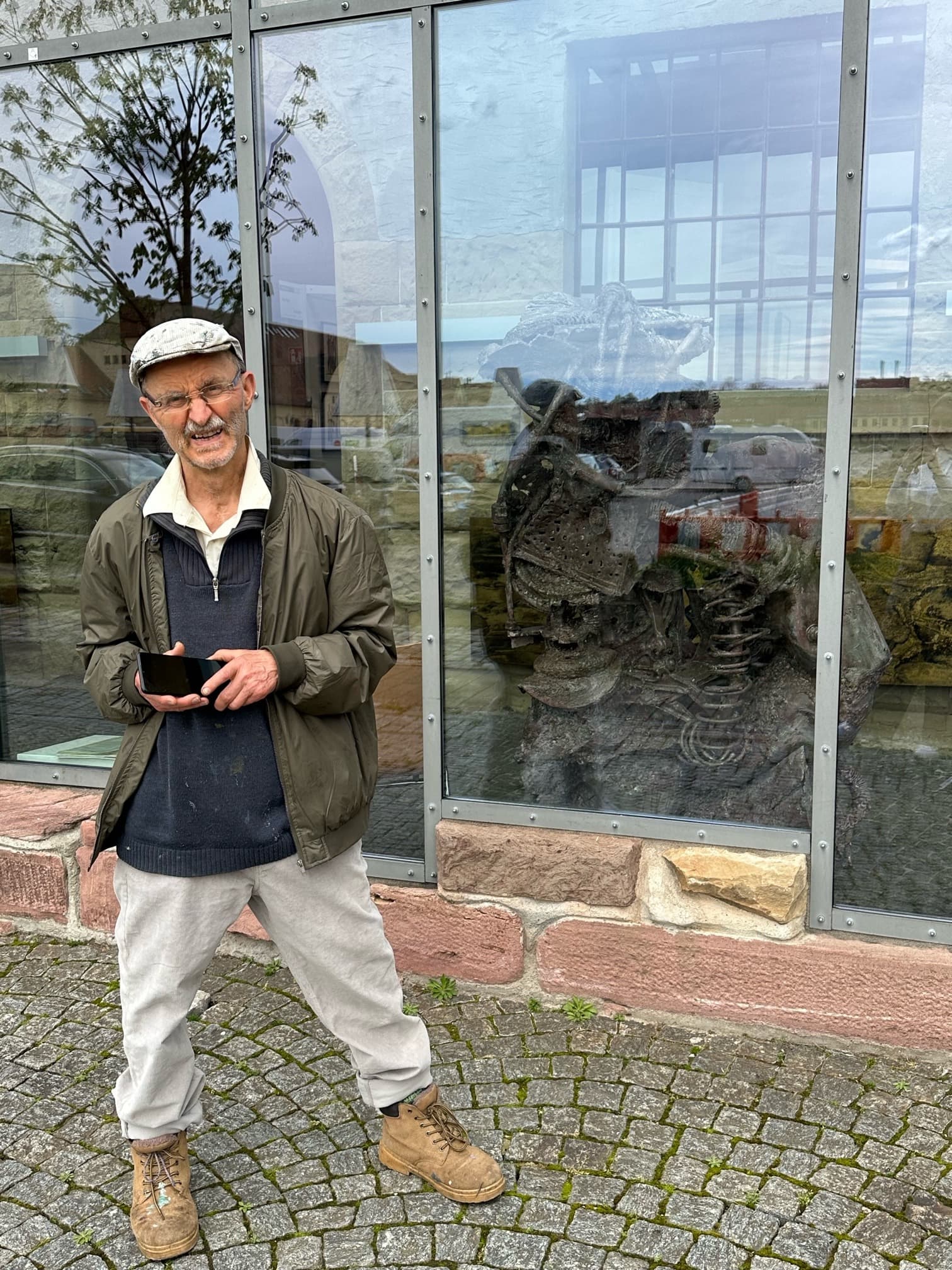 Gerd Georg Hirschmann mit Schiebermütze und grüner Jacke vor einer Glasfassade, hinter der Scheibe eine Metallskulptur