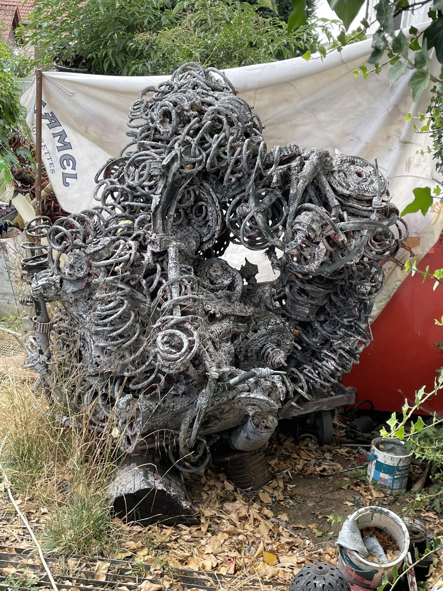 Silbergraue Metallskulptur aus Schlingen, Federn und Spiralen in einem verwilderten Garten vor weisser Plane