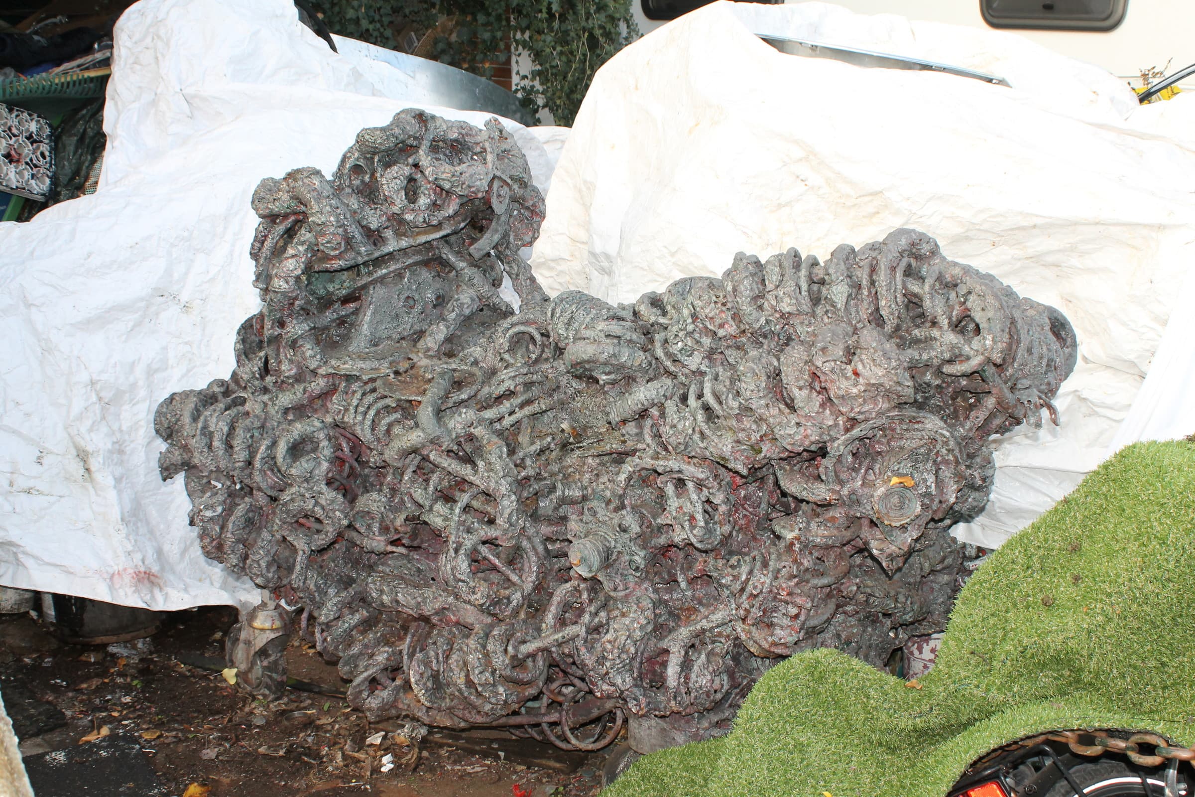 Breite grau-violette Metallskulptur aus verschweissten Schlingen und Federn, rote Grundierung scheint durch, auf weissen Planen und Kunstrasen