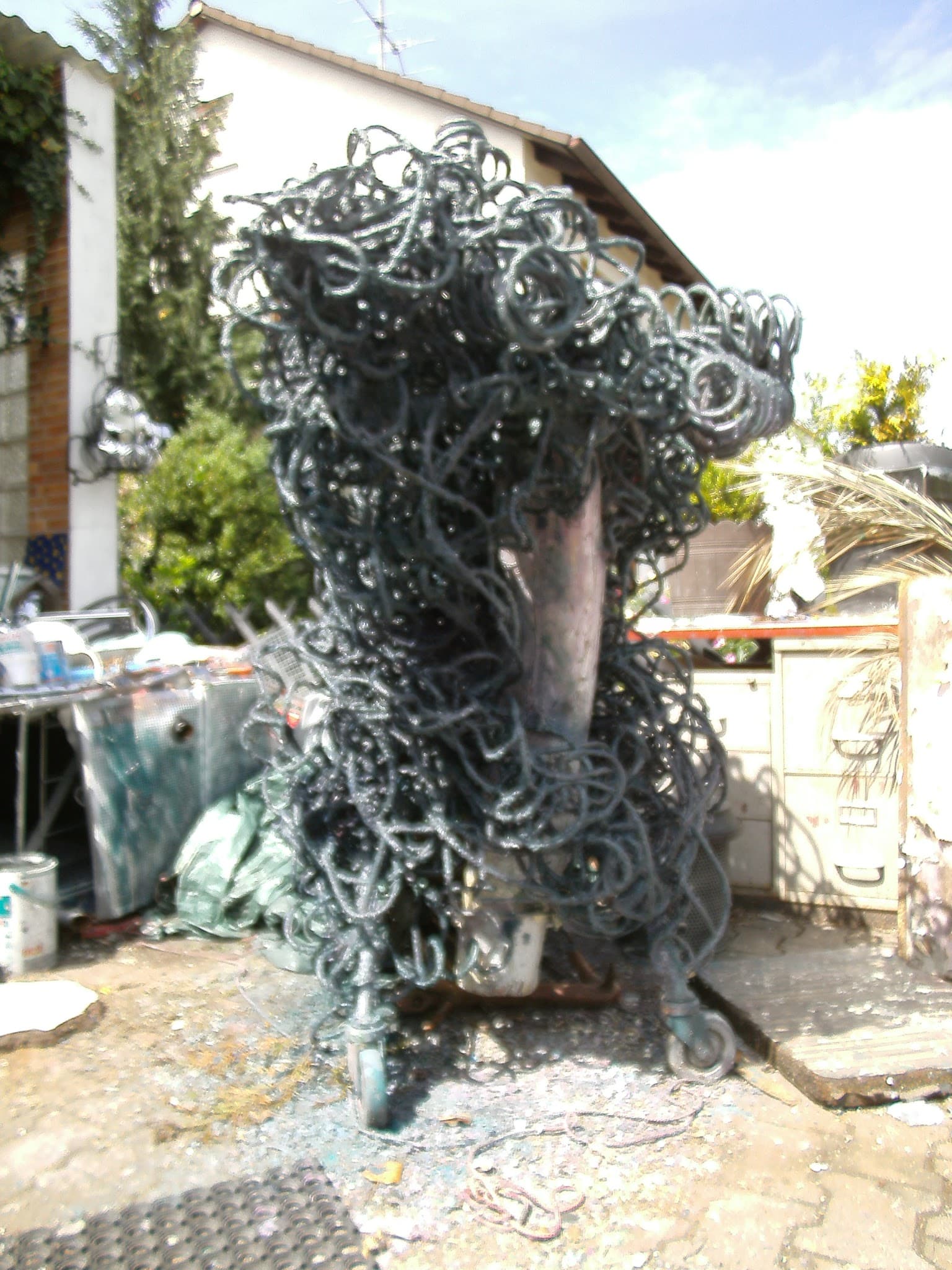 Hohe dunkel-grüngraue Schlingen-Skulptur auf Rollen mit kupfernem Oval in der Mitte, in einem Hof
