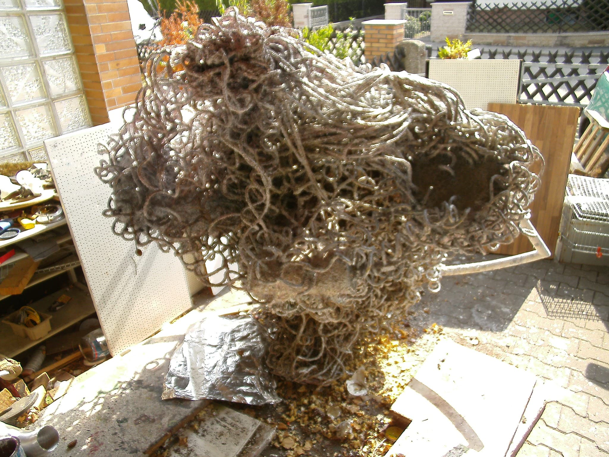 Hell-silbrige Schlingen-Skulptur in einem Hof vor einer Wand aus Glasbausteinen